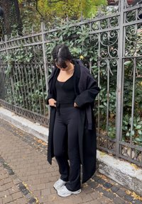 Manteau noir surdimensionné porté par-dessus un haut noir et un pantalon ajusté. Les chaussures ont un design épais avec des accents blancs. L'arrière-plan comprend un feuillage vert et une clôture en métal.