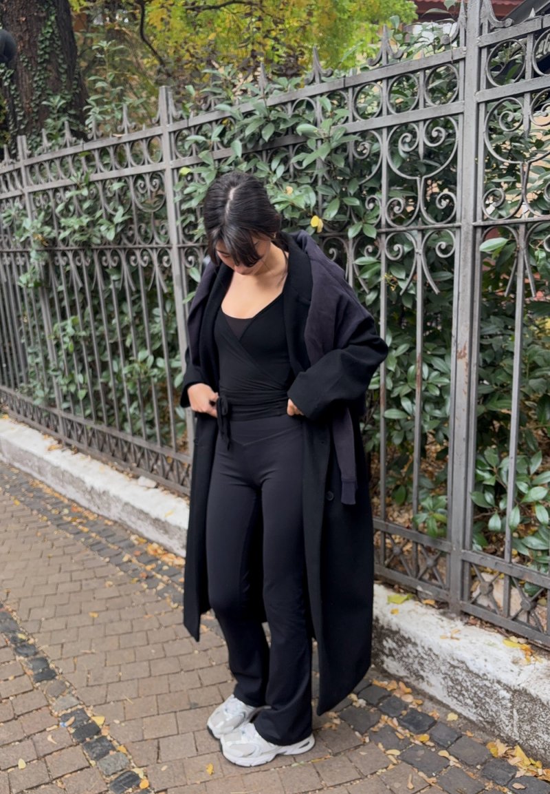 Manteau noir surdimensionné porté par-dessus un haut noir et un pantalon ajusté. Les chaussures ont un design épais avec des accents blancs. L'arrière-plan comprend un feuillage vert et une clôture en métal.