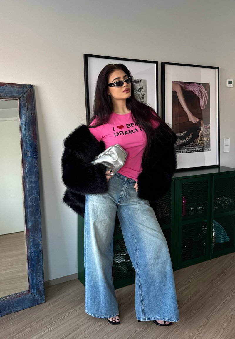 Vrouw met lang donker haar en zonnebril, roze T-shirt met grafische print, zwarte bontjas, wijde jeans, houdend een zilveren clutch, staand in een kamer met kunst en een spiegel.