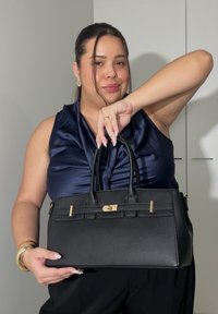 Femme en haut bleu marine tenant un sac à main en cuir noir avec ferrures dorées, debout devant un fond clair uni.