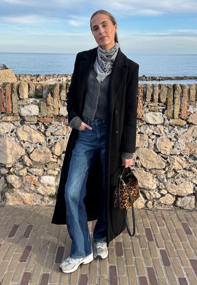 Joven mujer con abrigo largo negro, suéter gris, jeans azules, zapatillas blancas y pañuelo tipo bandana, de pie junto a un muro de piedra cerca del mar, sosteniendo un bolso de estampado de leopardo.