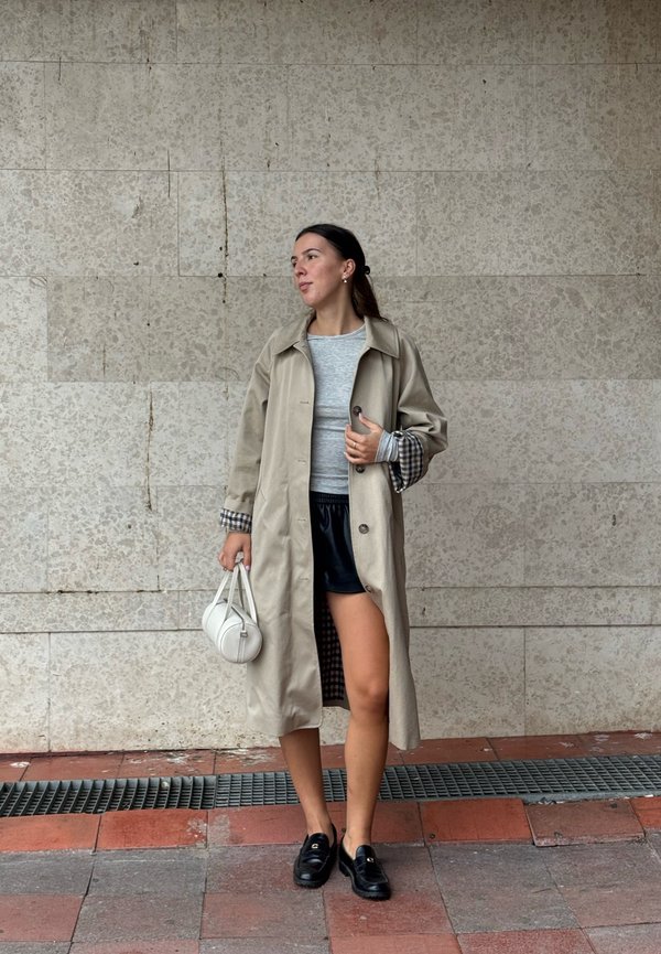 Una mujer con una gabardina beige, una camiseta gris, pantalones cortos negros y mocasines sostiene un bolso blanco mientras está junto a un muro de piedra.