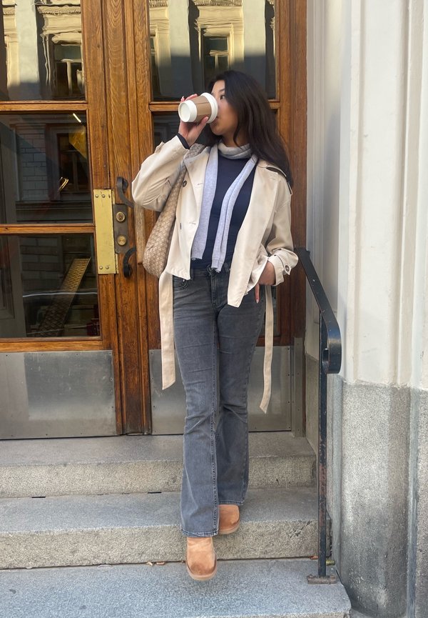 Gabardina beige, camiseta gris, bufanda gris claro, jeans acampanados grises y botas beige. Sosteniendo una taza de café, de pie en escalones de piedra cerca de puertas de madera.