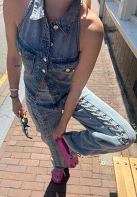 Denim jumpsuit featuring a halter neck design, light blue color, and lace-up sides; paired with bright pink sandals and sunglasses.