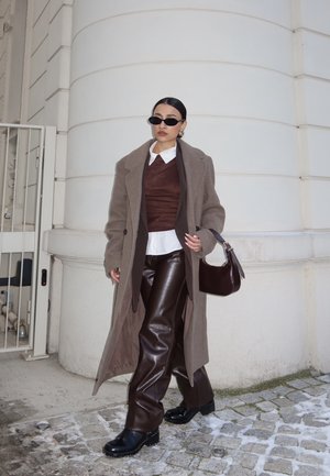 Mujer con abrigo largo marrón, pantalones de cuero, gafas de sol oscuras y sosteniendo un bolso oscuro, caminando por una calle empedrada cerca de un edificio blanco.