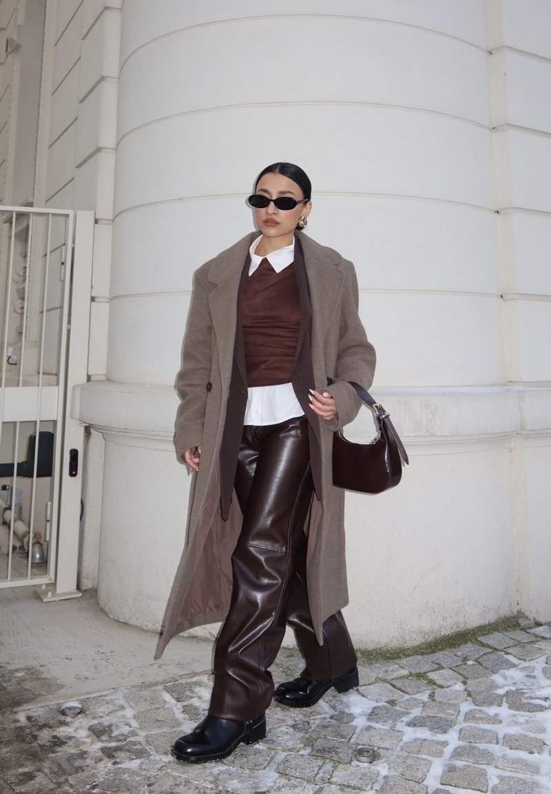 Femme portant un long manteau marron, un pantalon en cuir, des lunettes de soleil foncées, et tenant un sac à main foncé, marchant dans une rue pavée près d'un bâtiment blanc.