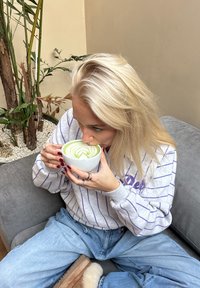 Bol en céramique blanche avec un art latte vert formant un motif tourbillonnant. La personne porte un sweat-shirt gris clair à rayures et est assise sur un canapé gris.