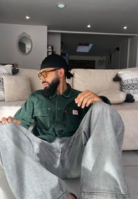 Grüne Cordhemd mit zwei Brusttaschen, graue, weite Jeans und gelb getönte Brille. Heller Sofa und strukturierte Kissen im Hintergrund.