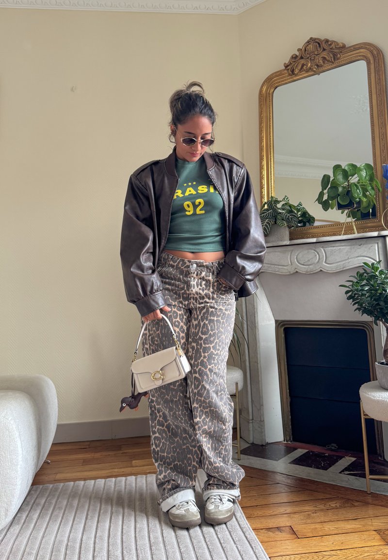 Brown leather jacket, green crop top with yellow text, leopard print trousers, white handbag, grey trainers, and sunglasses. Indoor setting.