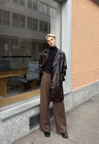 Jeune personne appuyée contre un mur de bâtiment, portant un manteau en cuir brun foncé, un col roulé noir, un pantalon large marron et des chaussures noires.