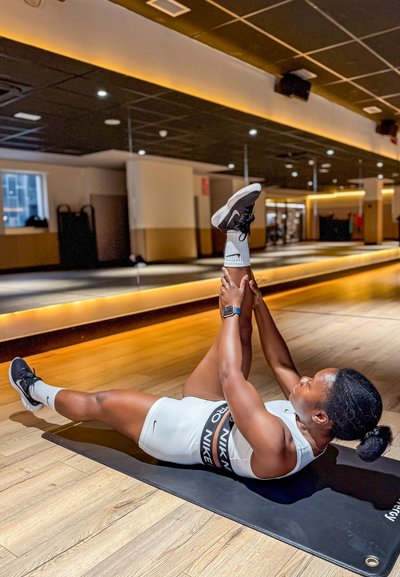 Persona estirándose en una esterilla de yoga negra en un estudio de fitness. Lleva un bodysuit blanco con un logo negro de Nike y zapatillas negras con calcetines blancos.