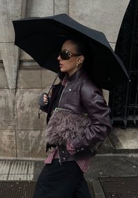 Veste en cuir couleur bordeaux profond, avec une pochette rehaussée de fourrure en laine. Parapluie noir tenu au-dessus, associé à des lunettes de soleil foncées et une chemise rayée.
