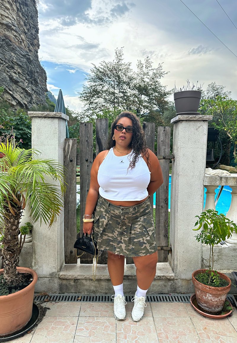 Top court blanc associé à une mini-jupe camouflage. Les accessoires comprennent un petit sac à main noir et des chaussures de sport blanches. Cadre extérieur.