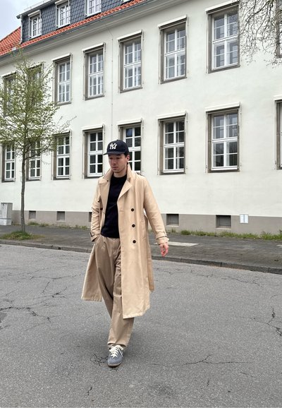 Hombre caminando por una calle vestido con un abrigo beige, camisa azul marino, pantalones beige, zapatillas grises y una gorra de béisbol negra con letras blancas.