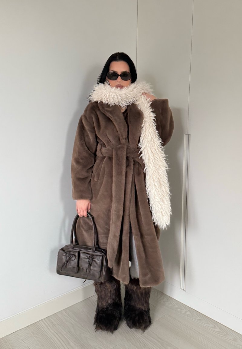Manteau marron en fausse fourrure avec ceinture, associé à une longue écharpe blanche texturée et un sac en cuir noir. Détails en fausse fourrure noire sur les jambes.