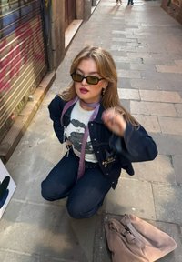 Τζάκετ από τζιν πάνω από ένα γραφικό t-shirt, σκούρα τζιν, και γυαλιά ηλίου; ροζ κασκόλ δεμένο στον λαιμό; ανοιχτό καφέ τσάντα στο έδαφος.