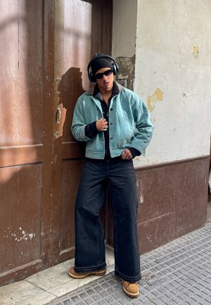 Chaqueta de gran tamaño azul claro con cuello negro, llevada sobre una camiseta negra. Jeans oscuros de corte holgado y botines color beige. Auriculares negros y gafas de sol.