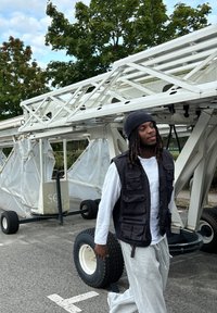 Μαύρο γιλέκο με πολλές τσέπες, φορεμένο πάνω από λευκό πουκάμισο με μακριά μανίκια, σε συνδυασμό με γκρι φόρμες. Μεγάλος εξοπλισμός στο βάθος.