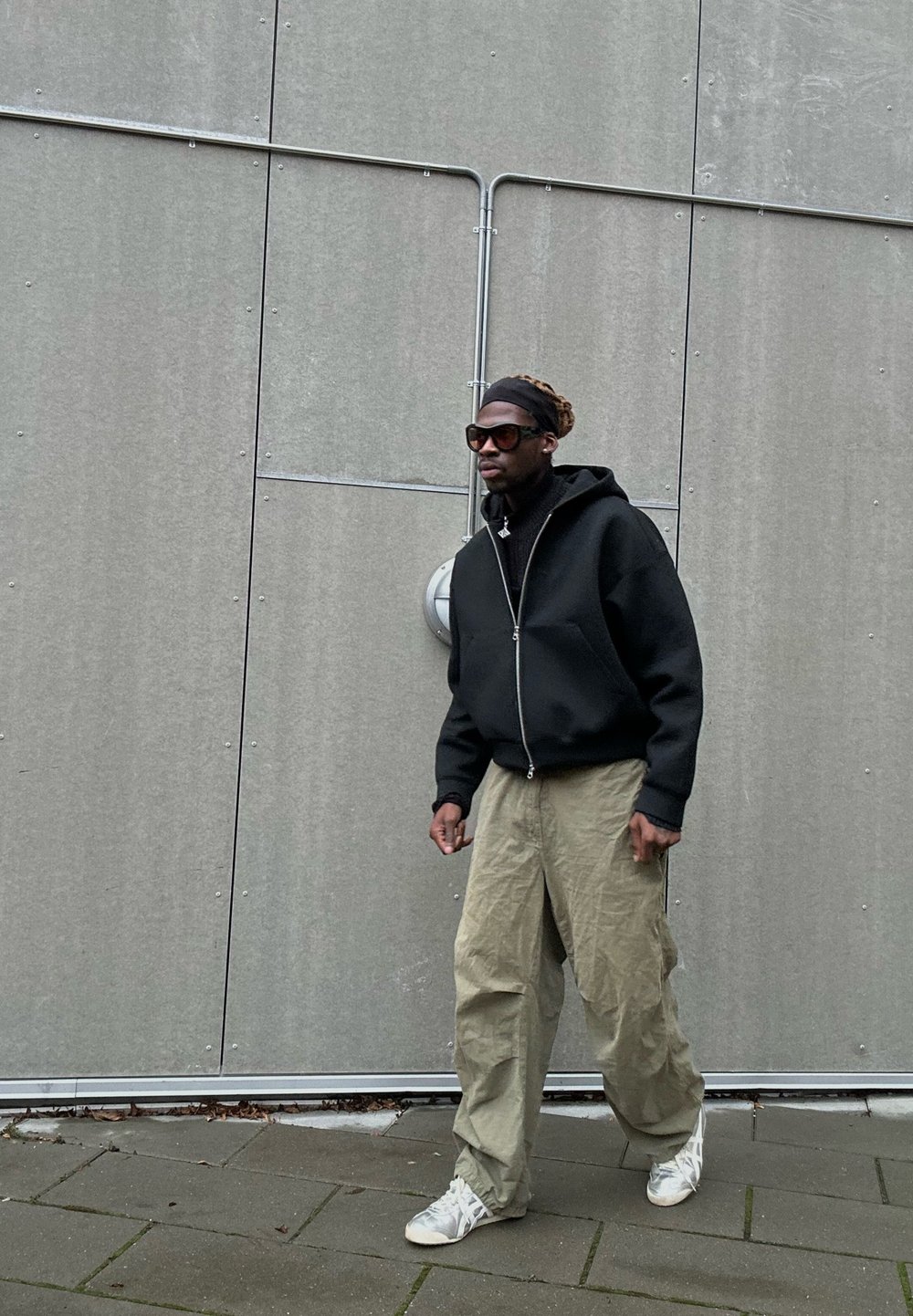 Hoodie nero con zip, pantaloni larghi verde oliva, scarpe sportive bianche, occhiali da sole grandi, in piedi contro un muro grigio con dettagli in tubo.