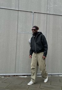 Hoodie nero con zip, pantaloni larghi verde oliva, scarpe sportive bianche, occhiali da sole grandi, in piedi contro un muro grigio con dettagli in tubo.