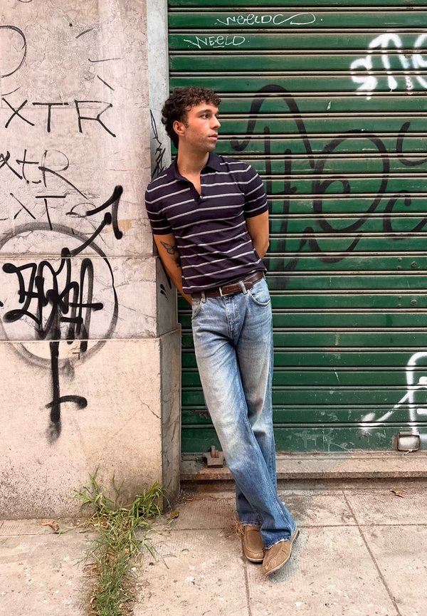 Young man in striped shirt and jeans leans against graffiti-covered wall and green metal shutter, looking to his left.