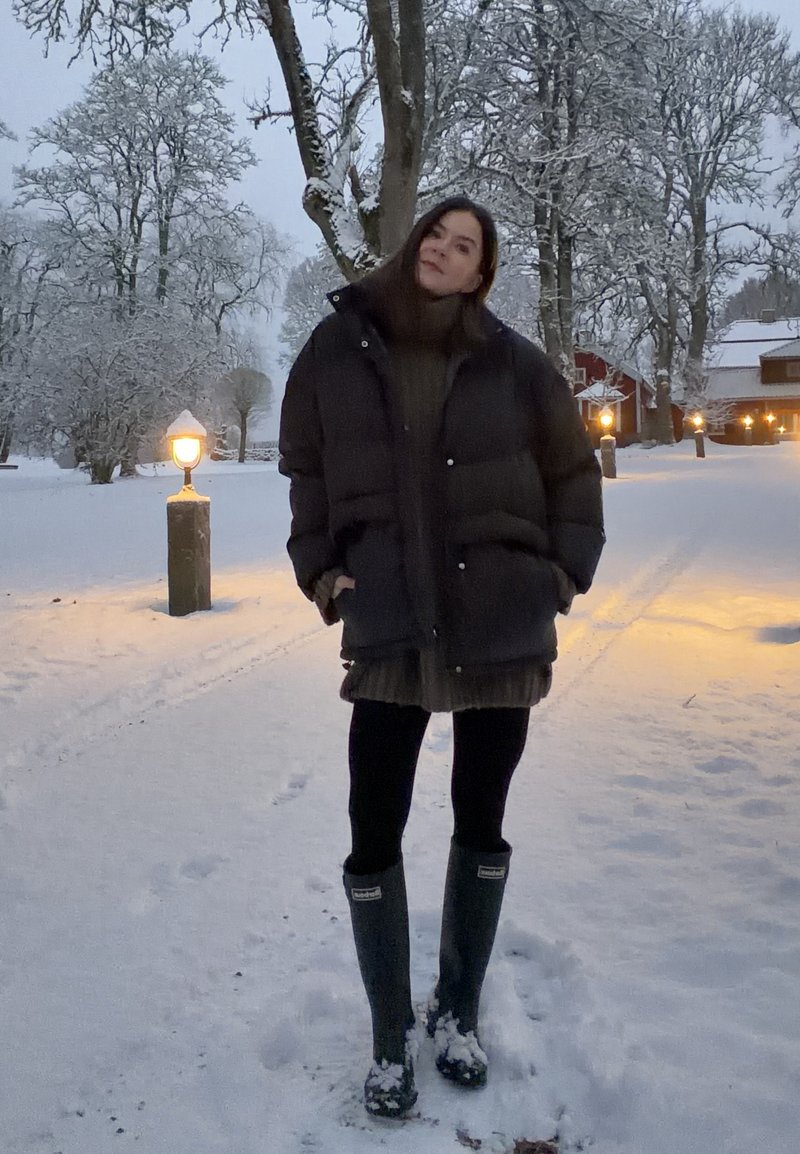 Lockere dunkle Jacke, gerippten dunklen Pullover, mehrlagigen Rock, schwarze Leggings und grüne Gummistiefel. Verschneite Landschaft mit Straßenlaternen und Bäumen.