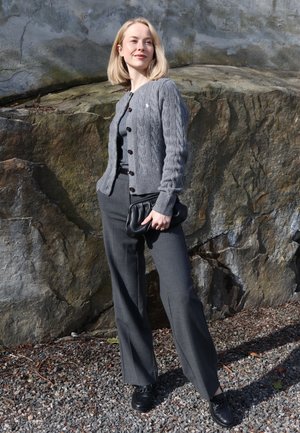 Mujer con cárdigan de punto gris, pantalones a juego y zapatos negros, de pie al aire libre frente a una gran pared de roca, sosteniendo un bolso de mano negro.