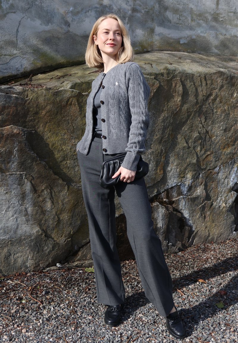 Femme en cardigan tricoté gris, pantalon assorti et chaussures noires, debout à l'extérieur contre un grand mur de roche, tenant une pochette noire.