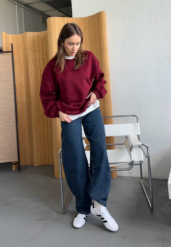 Sweatshirt oversize bordeaux, chemise blanche en superposition, jeans larges bleu foncé et baskets blanches avec des rayures noires, sur un fond neutre.