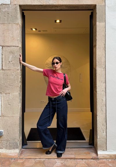 Mujer con gafas de sol, que lleva una camisa rosa y vaqueros de pierna ancha, de pie en una puerta de piedra con un brazo extendido y un bolso negro en el hombro.