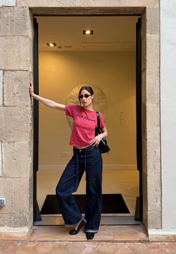Frau mit Sonnenbrille, die ein rosa Hemd und Jeans mit weitem Bein trägt, steht in einem steinernen Türrahmen, mit einem ausgestreckten Arm und einer schwarzen Tasche auf der Schulter.