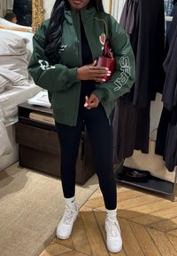 Persona con chaqueta verde oscura con logotipos, leggings negros, zapatillas blancas Nike, sosteniendo un pequeño bolso rojo en una habitación con ropa y muebles.