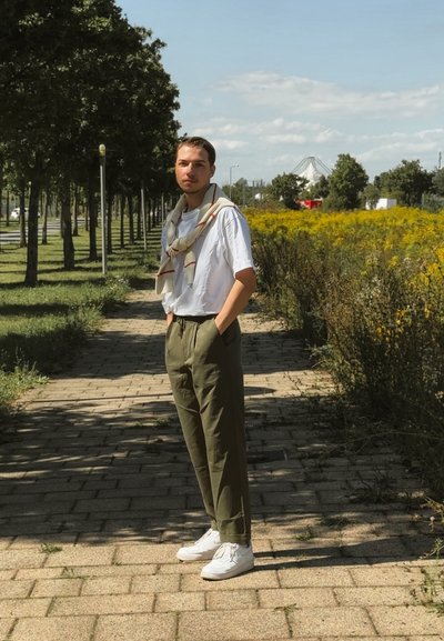 Persona con una camisa blanca de manga corta, pantalones verde oliva y zapatillas blancas, con una bufanda con estampado drapeada alrededor del cuello.