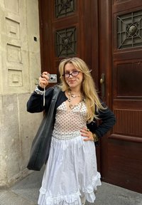 Una persona tiene in mano una macchina fotografica compatta, indossa un top a pois e una gonna bianca a volant, abbinati a un cardigan nero e a una grande borsa nera.