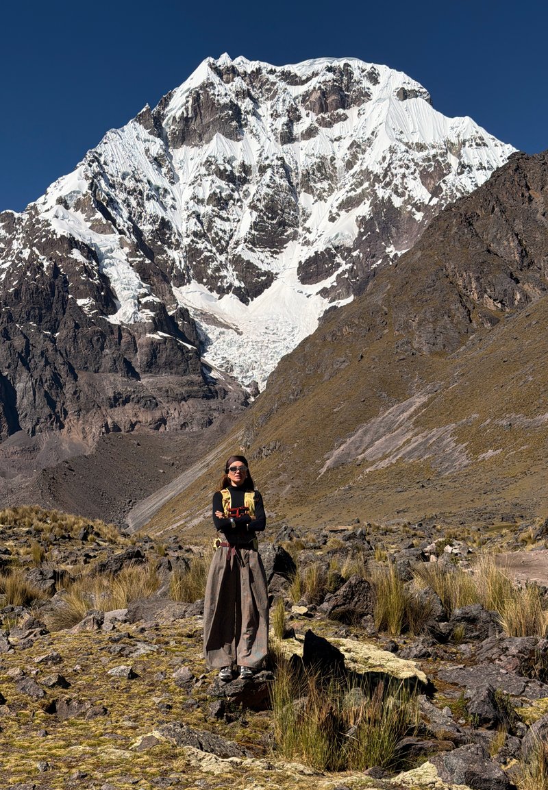 Femme en tenue d'extérieur debout sur un sol rocheux avec de hautes herbes devant une grande montagne enneigée sous un ciel bleu clair.