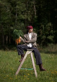 Personne assise sur une échelle en bois dans un champ d'herbe, tenant un bouquet de carottes fraîches, portant un bandana rouge, un blazer et des bottes.