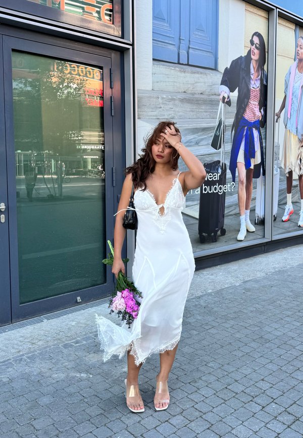 Femme en robe nuisette blanche en dentelle tenant des fleurs roses, debout sur le trottoir devant des vitrines avec des affiches de mode.