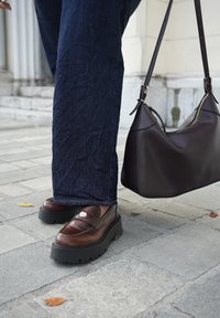 Mocassins en cuir marron avec semelles épaisses noires et un accent argenté. Jeans bleu foncé avec une finition texturée. Un sac à main en cuir marron foncé.