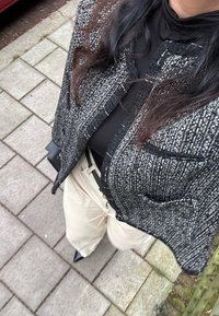 Črna in bela strukturalna tweed jakna z raztrganimi robovi, oblečena čez pulover s turtleneck izrezom, v kombinaciji s bež hlačami in črnimi podpetimi čevlji.