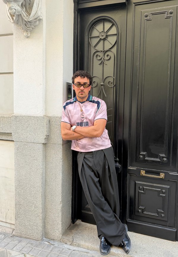Homme portant des lunettes de soleil, un t-shirt Puma rose, un pantalon large noir et des chaussures noires s'appuyant contre un mur de pierre gris près d'une porte noire décorative.