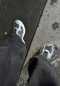 Sportskor i grått mesh med svarta accenter och reflekterande detaljer. Bärs med löst sittande svarta byxor, placerade på en strukturerad asfalt.