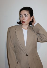 Femme aux cheveux courts et foncés, portant un blazer beige oversize et une chemise blanche, posant avec la main touchant ses cheveux contre un mur uni.