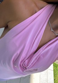 Femme portant un haut sans manches rose clair drapé et un collier argenté, debout près d'un sol en carrelage clair et d'une pelouse verte.