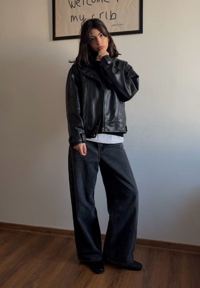 Chaqueta de cuero negra con solapas anchas, combinada con una camiseta negra y jeans sueltos de color gris oscuro. Botas negras de tacón completan el conjunto.