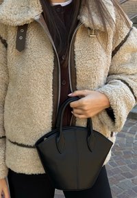 Jachetă bej din shearling cu margini din piele maro, asortată cu o geantă neagră structurată, care evidențiază o textură netedă și o siluetă rotunjită.