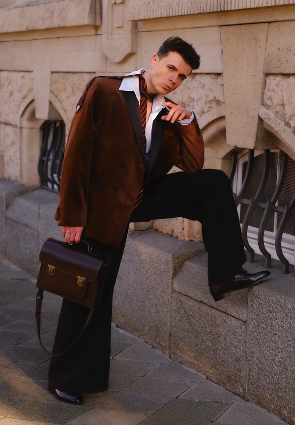 Blazer doppiopetto in velluto marrone con revers neri, camicia bianca, cravatta a righe, pantaloni neri e scarpe lucide. Borsa in pelle marrone.