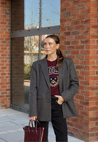 Blazer cinza sob medida sobre um suéter bordô com um emblema gráfico, combinado com jeans pretos e uma bolsa em relevo de crocodilo bordô.