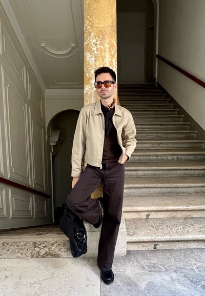 Chaqueta beige, camisa negra, pantalones marrones, zapatos negros. Gafas de sol marrones, una bufanda de estampado, de pie junto a una columna de mármol en las escaleras.