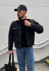 Veste noire matelassée avec un col en fausse peau de mouton beige, portée sur un pull noir et un jean bleu. Tient un sac fourre-tout noir.