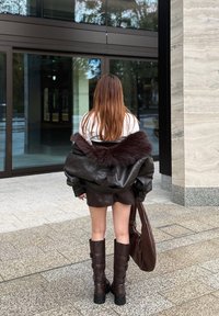 Smeđa kožna jakna s ovratnikom od umjetnog krzna, uparena sa smeđom kožnom mini suknjom i čizmama do koljena s kopčama, te torbom u istom stilu.